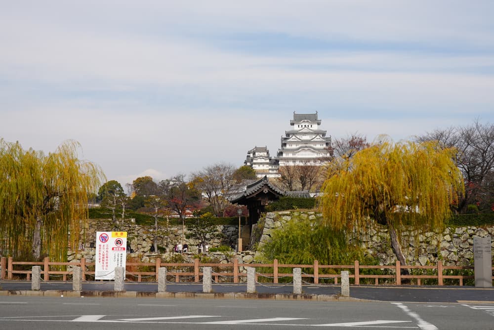 Himeji Castle, white against a pale sky, behind willows and a stone wall